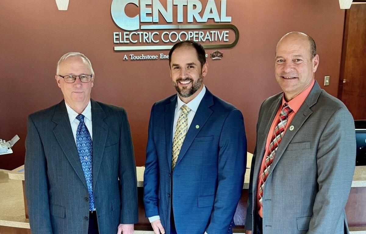 Three men in business suits posing for a photo in front of a Central Electric Cooperative sign in a lobby.