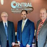 Three men in business suits posing for a photo in front of a Central Electric Cooperative sign in a lobby.