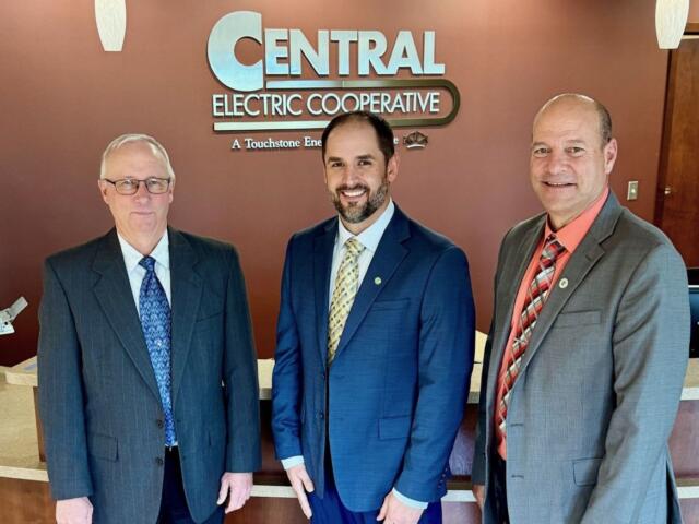 Three men in business suits posing for a photo in front of a Central Electric Cooperative sign in a lobby.