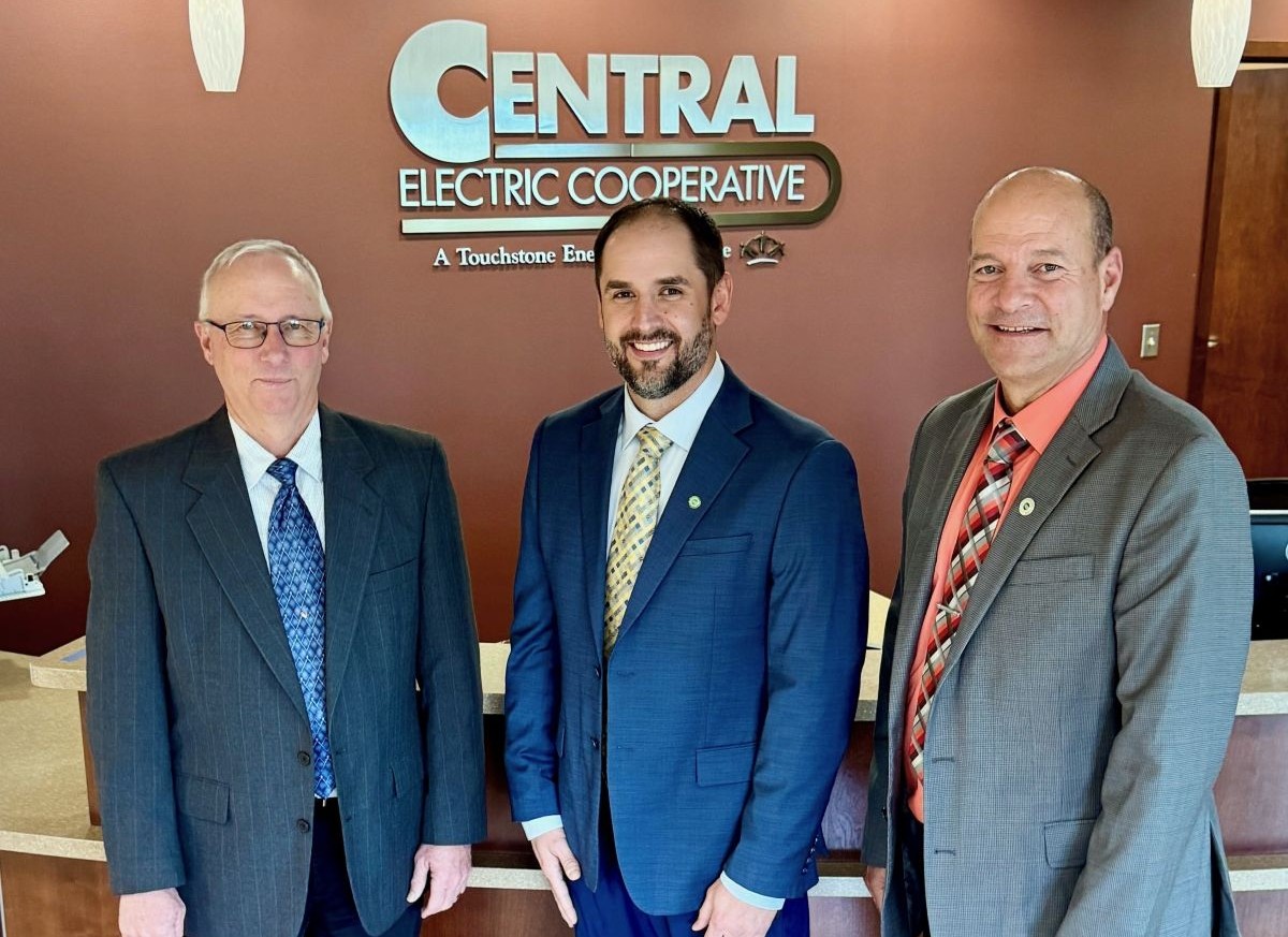 Three men in business suits posing for a photo in front of a Central Electric Cooperative sign in a lobby.