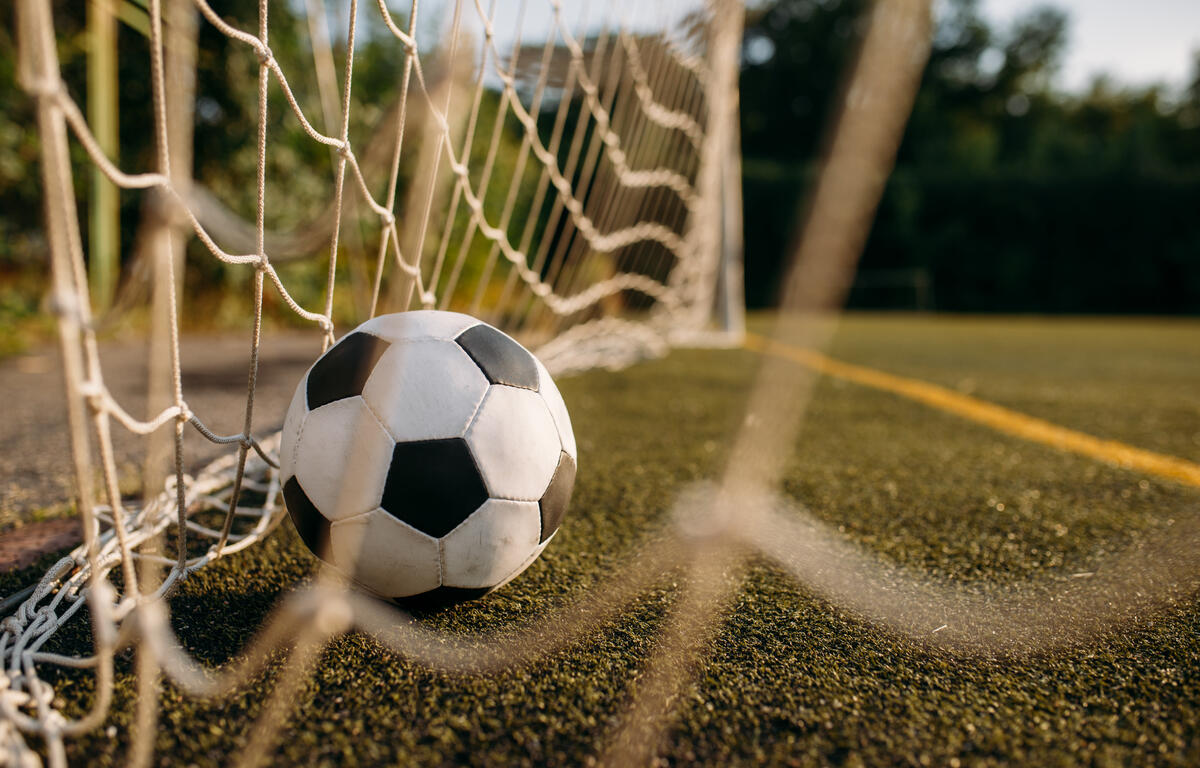 Soccer ball resting against a goal net on a grass/astro turf field, close-up view from ground level.