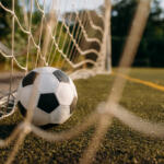 Soccer ball resting against a goal net on a grass/astro turf field, close-up view from ground level.