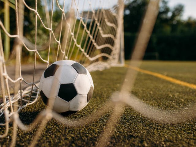 Soccer ball resting against a goal net on a grass/astro turf field, close-up view from ground level.