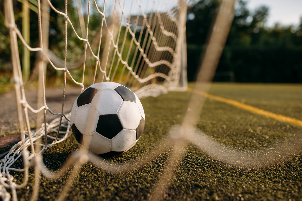 Soccer ball resting against a goal net on a grass/astro turf field, close-up view from ground level.