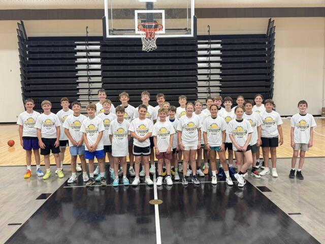 Group of young basketball players in white Kernel camp shirts posing for a group photo in the Mitchell high school main gym