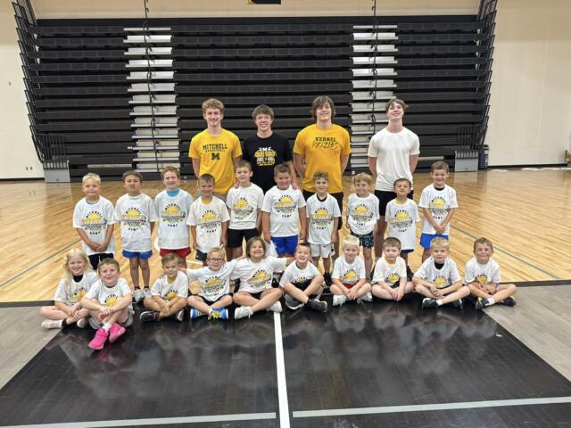 Group of young basketball players in white Kernel camp shirts posing for a group photo in the Mitchell high school main gym with current Mitchell varsity basketball players