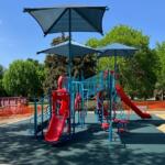 Colorful playground structure with red slides and blue climbing frames under large shade umbrellas in a park on a sunny day.