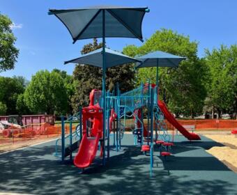 Colorful playground structure with red slides and blue climbing frames under large shade umbrellas in a park on a sunny day.