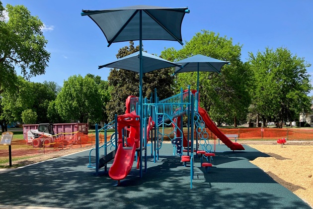 Colorful playground structure with red slides and blue climbing frames under large shade umbrellas in a park on a sunny day.