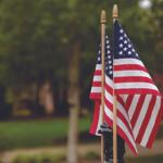 Two American flags on wooden poles waving in a park breeze, with a blurred green background.