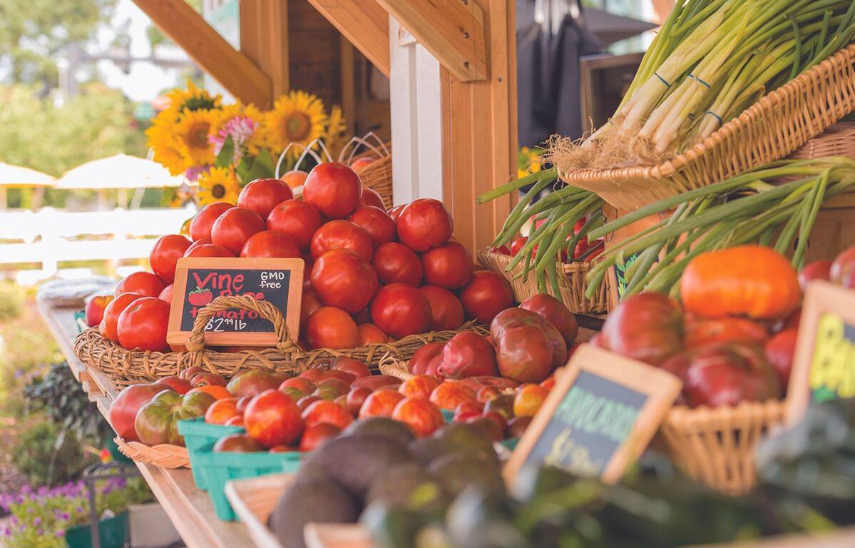 Stack of ripe red tomatoes in wicker baskets at an outdoor farmer's market, with leeks in a basket to the right and sunflowers in the background.