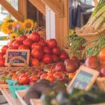 Stack of ripe red tomatoes in wicker baskets at an outdoor farmer's market, with leeks in a basket to the right and sunflowers in the background.