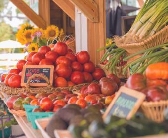Stack of ripe red tomatoes in wicker baskets at an outdoor farmer's market, with leeks in a basket to the right and sunflowers in the background.