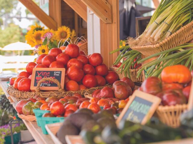 Stack of ripe red tomatoes in wicker baskets at an outdoor farmer's market, with leeks in a basket to the right and sunflowers in the background.