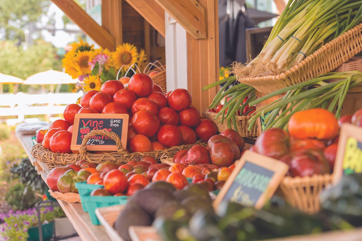 Stack of ripe red tomatoes in wicker baskets at an outdoor farmer's market, with leeks in a basket to the right and sunflowers in the background.