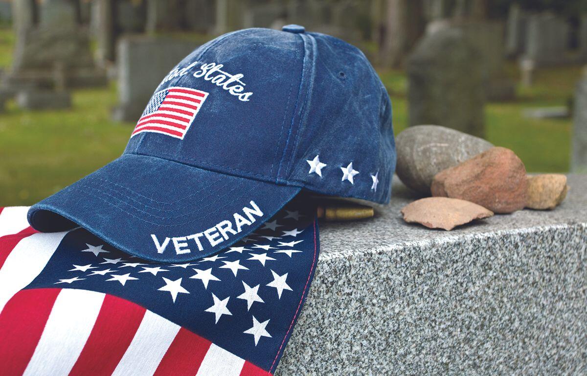 Patriotic blue veteran cap with US flag patch rests on a granite memorial beside scattered stones in a cemetery.