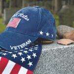 Patriotic blue veteran cap with US flag patch rests on a granite memorial beside scattered stones in a cemetery.
