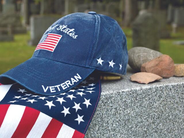 Patriotic blue veteran cap with US flag patch rests on a granite memorial beside scattered stones in a cemetery.