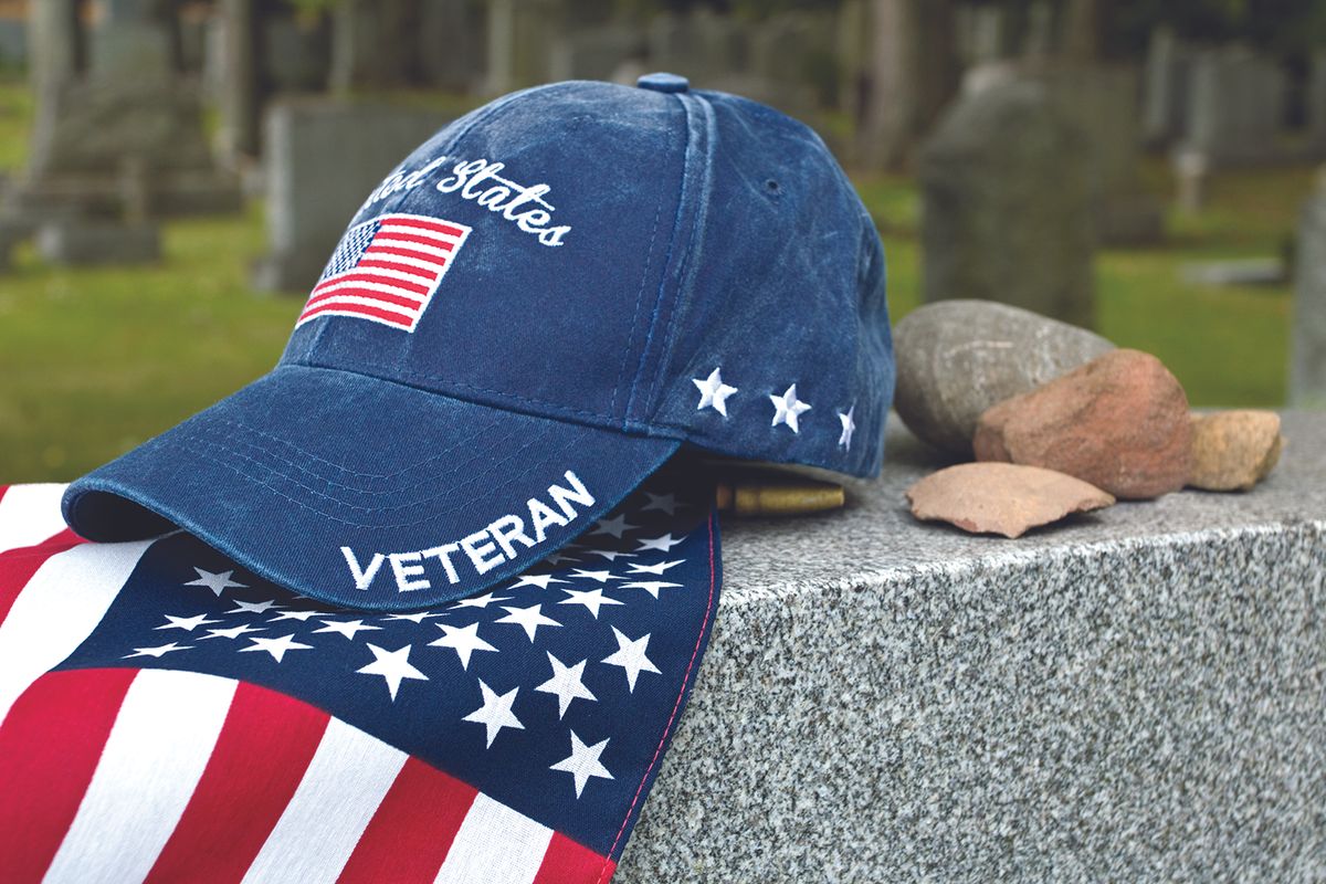 Patriotic blue veteran cap with US flag patch rests on a granite memorial beside scattered stones in a cemetery.