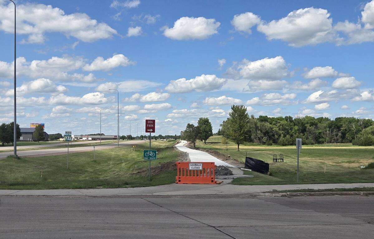 Construction barrier on a sidewalk with a 'Sidewalk Closed' sign; a new path under construction runs through a grassy area toward trees under a blue sky with clouds.
