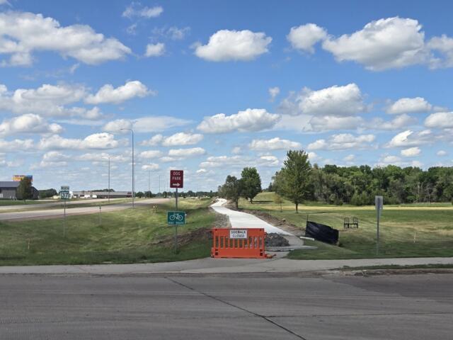 Construction barrier on a sidewalk with a 'Sidewalk Closed' sign; a new path under construction runs through a grassy area toward trees under a blue sky with clouds.