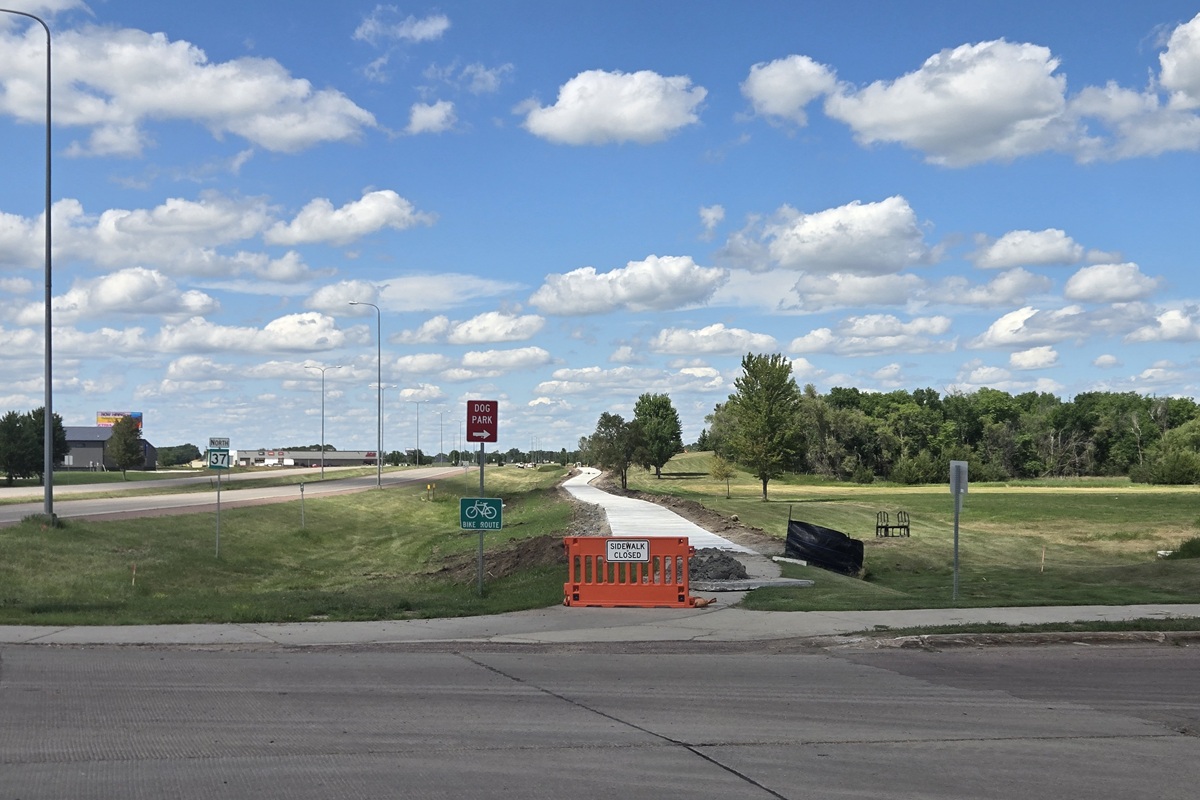 Construction barrier on a sidewalk with a 'Sidewalk Closed' sign; a new path under construction runs through a grassy area toward trees under a blue sky with clouds.