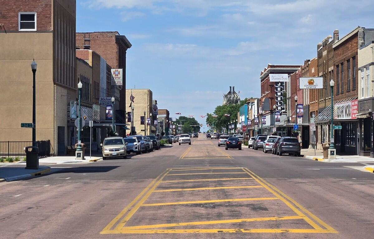 Downtown main street lined with brick storefronts and parked cars on both sides, yellow crosswalk markings in the foreground under a blue sky.