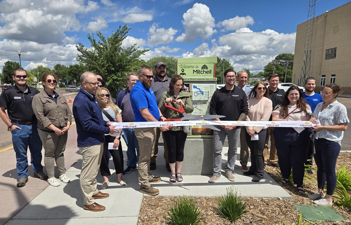 Group of people at a ribbon-cutting ceremony outdoors, cutting a white ribbon in front of a Welcome to Mitchell, South Dakota sign.