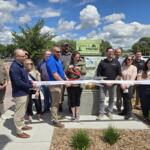 Group of people at a ribbon-cutting ceremony outdoors, cutting a white ribbon in front of a Welcome to Mitchell, South Dakota sign.