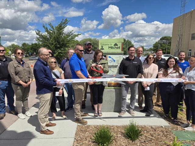 Group of people at a ribbon-cutting ceremony outdoors, cutting a white ribbon in front of a Welcome to Mitchell, South Dakota sign.
