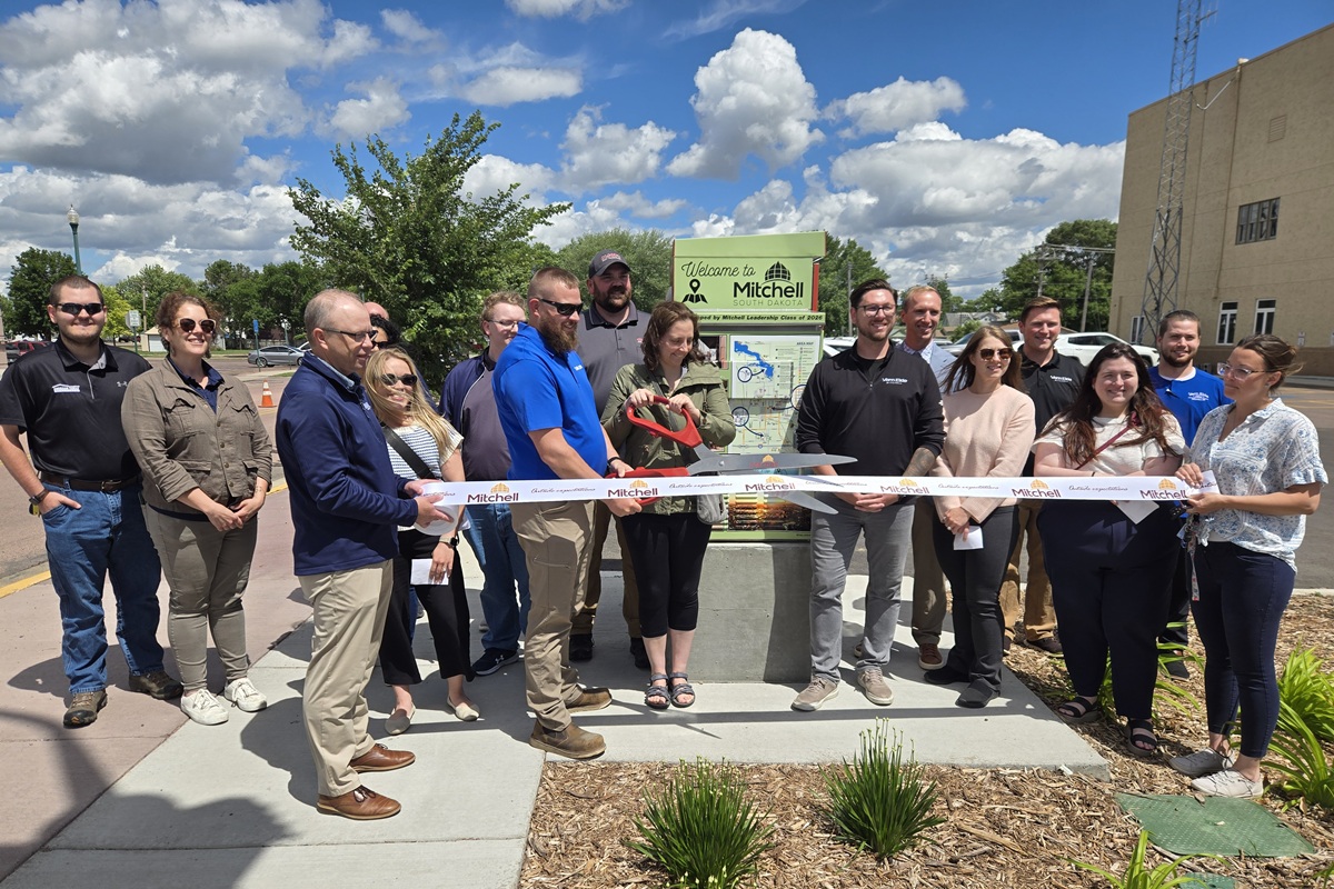 Group of people at a ribbon-cutting ceremony outdoors, cutting a white ribbon in front of a Welcome to Mitchell, South Dakota sign.