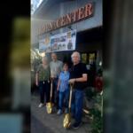 Four adults pose outside a garden center entrance, each holding a gold-colored shovel with a banner of photos above them.