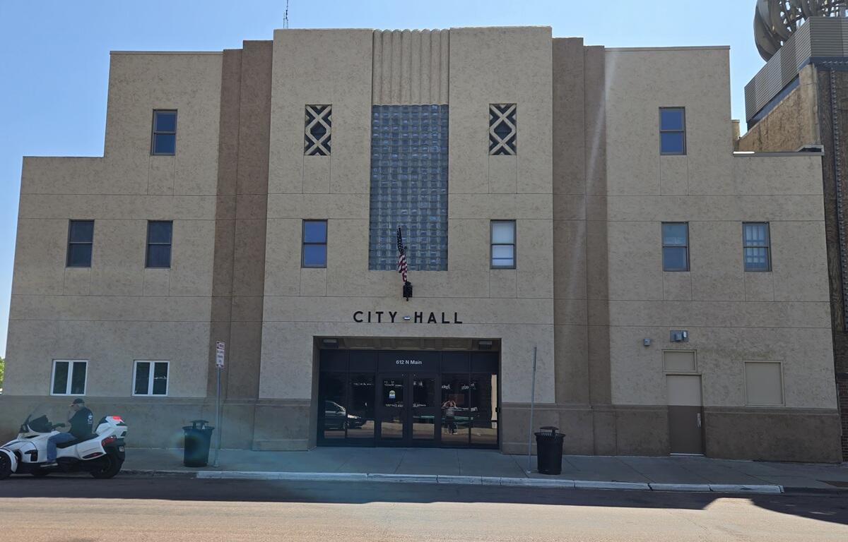 Front of City Hall with a beige facade, central glass-block feature, flag hanging by the entrance, and double doors below. (Informative)