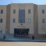 Front of City Hall with a beige facade, central glass-block feature, flag hanging by the entrance, and double doors below. (Informative)