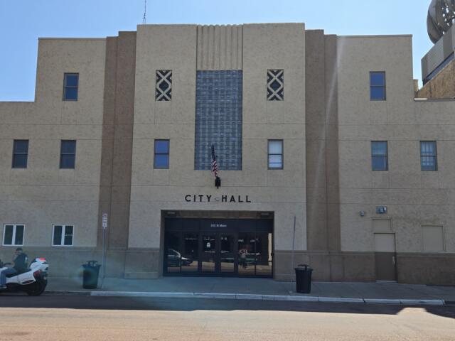 Front of City Hall with a beige facade, central glass-block feature, flag hanging by the entrance, and double doors below. (Informative)