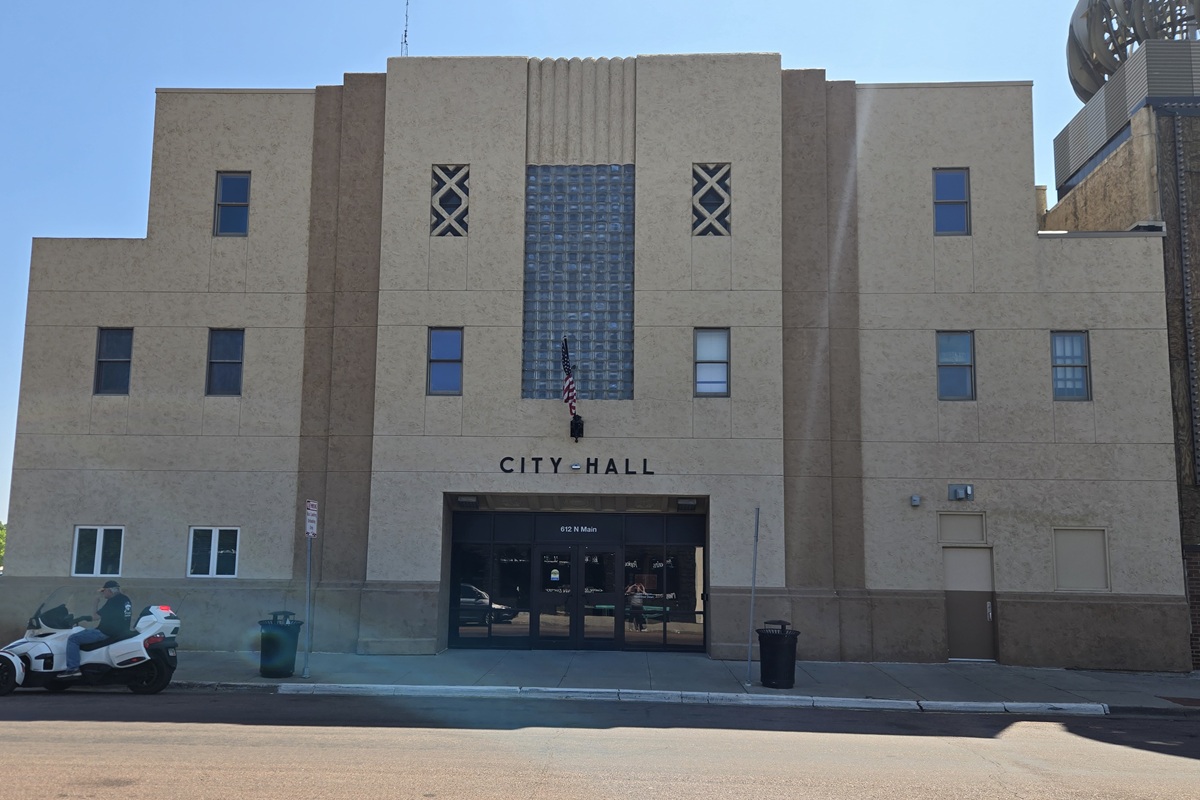 Front of City Hall with a beige facade, central glass-block feature, flag hanging by the entrance, and double doors below. (Informative)