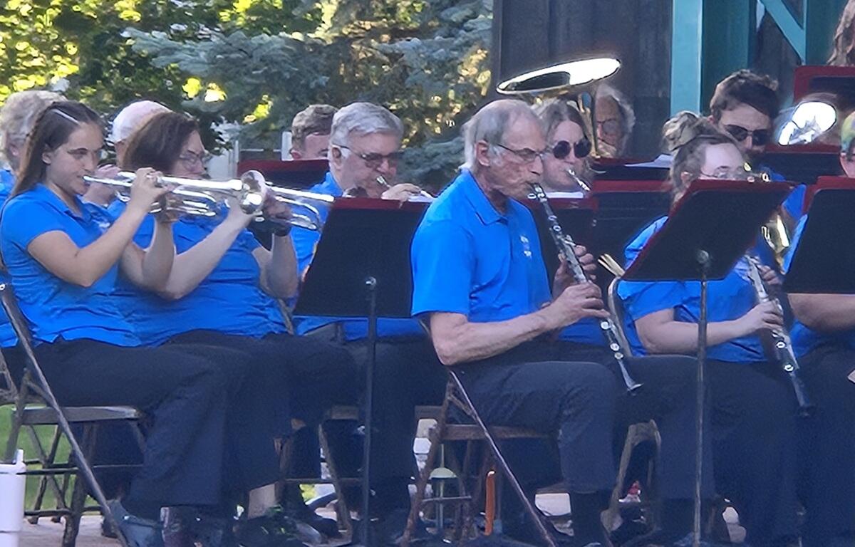 Outdoor community band performing in blue shirts; trumpet players on the left and a clarinetist center with music stands nearby.