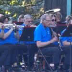 Outdoor community band performing in blue shirts; trumpet players on the left and a clarinetist center with music stands nearby.
