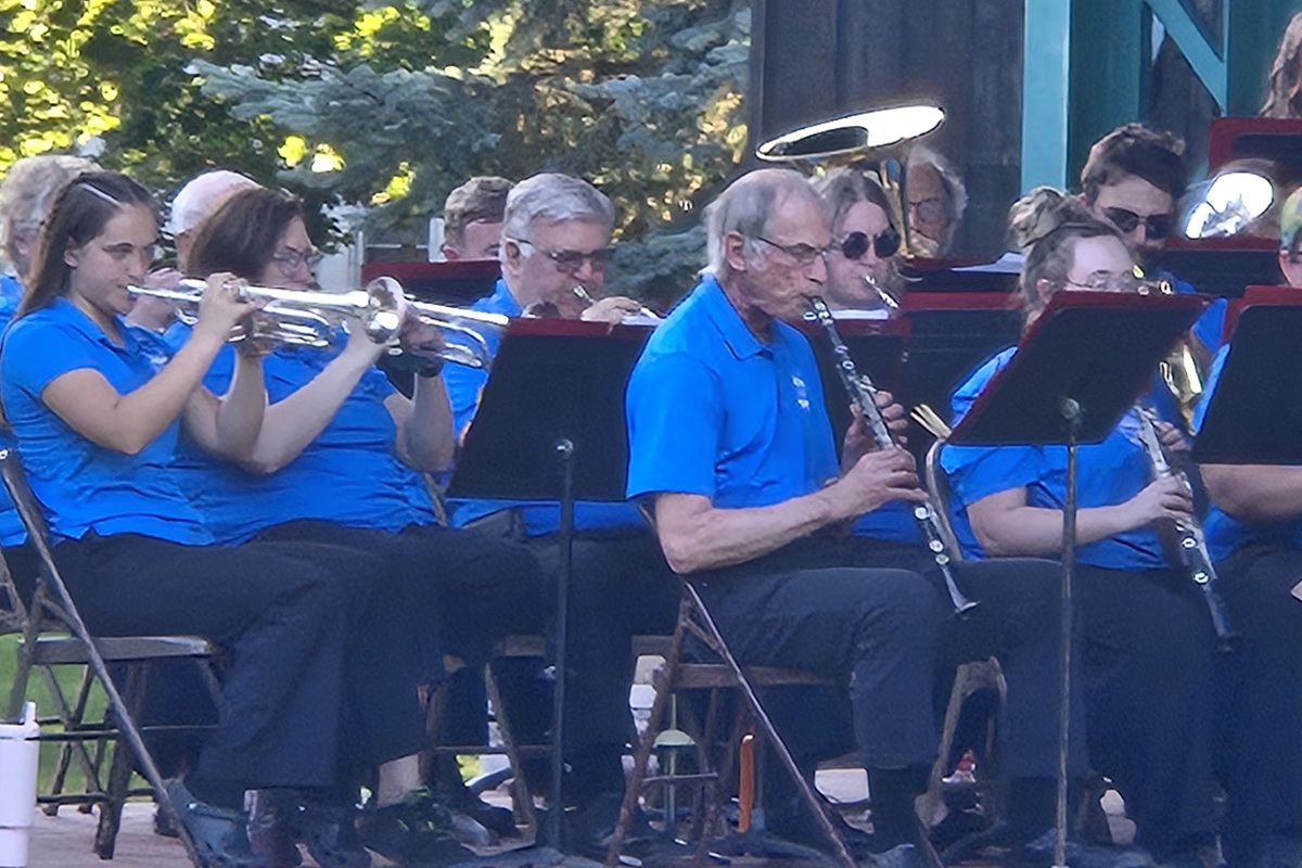 Outdoor community band performing in blue shirts; trumpet players on the left and a clarinetist center with music stands nearby.