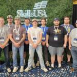 Group of nine men posing for a photo at a SkillUSA event, wearing lanyards and badges in front of a greenscreen backdrop with SkillUSA logo.