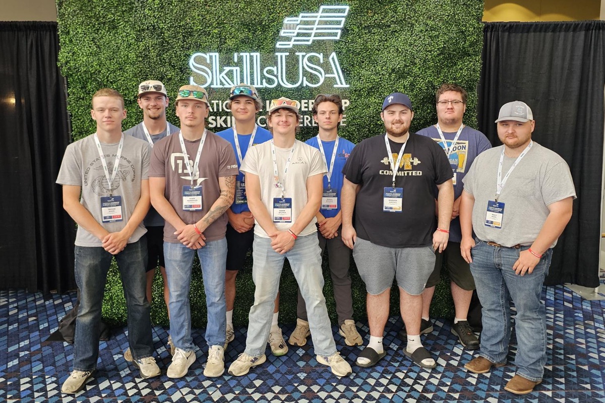 Group of nine men posing for a photo at a SkillUSA event, wearing lanyards and badges in front of a greenscreen backdrop with SkillUSA logo.