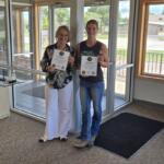Two women stand in a lobby holding certificates, smiling at the camera.