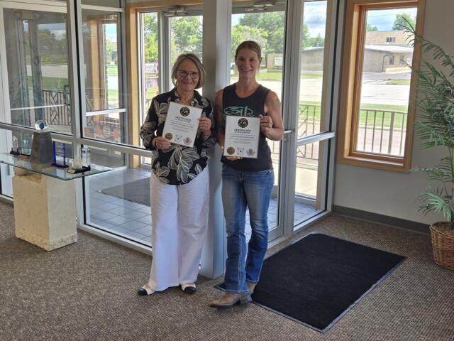 Two women stand in a lobby holding certificates, smiling at the camera.