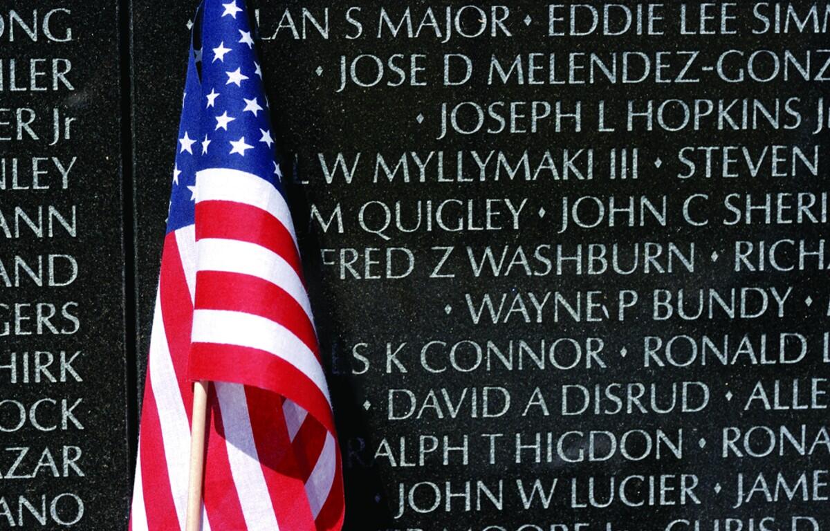 American flag draped in front of a memorial wall engraved with names on dark granite surface, commemorating the fallen.
