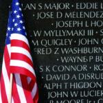 American flag draped in front of a memorial wall engraved with names on dark granite surface, commemorating the fallen.