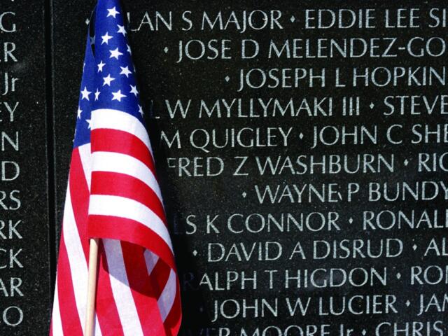 American flag draped in front of a memorial wall engraved with names on dark granite surface, commemorating the fallen.