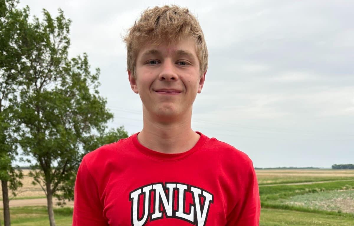 Teenager wearing a red UNLV shirt stands outdoors with trees and fields in the background, smiling at the camera.
