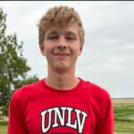Teenager wearing a red UNLV shirt stands outdoors with trees and fields in the background, smiling at the camera.