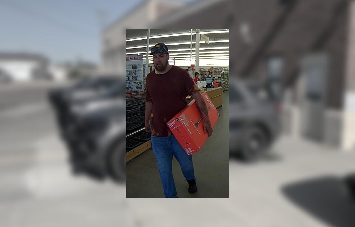 Bearded man in a burgundy shirt and cap carries a red-orange box through a store aisle.