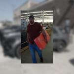 Bearded man in a burgundy shirt and cap carries a red-orange box through a store aisle.